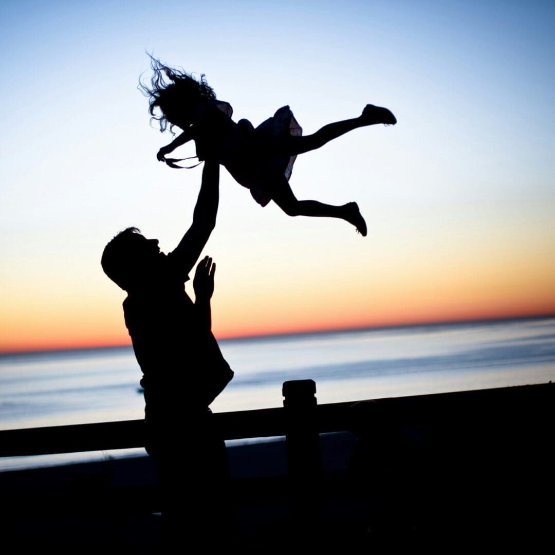 Adult lifting a child into the air at sunset, representing trust, joy, and healing in co-parenting therapy.