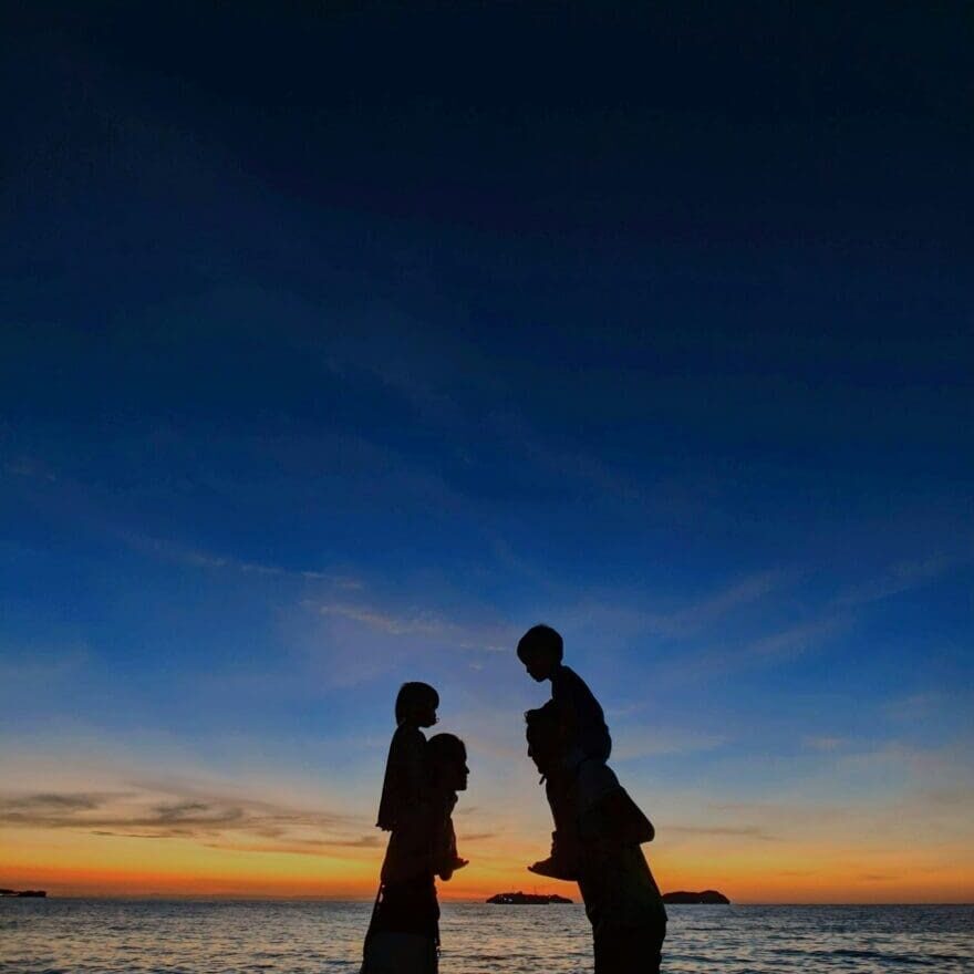 Two adults carrying children on their shoulders by the water at dusk, symbolizing stability and connection in co-parenting therapy.